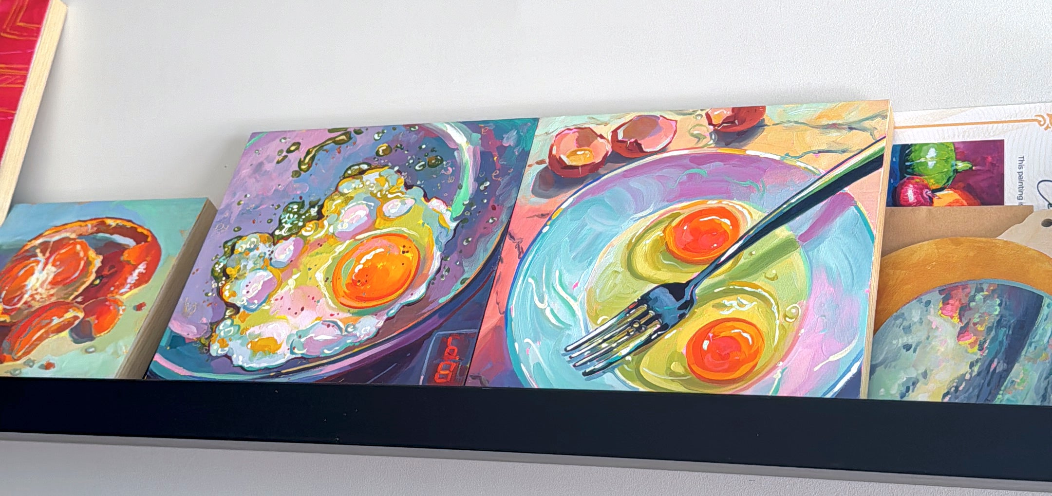 Colorful still life oil paintings by Alai Ganuza displayed on a black shelf, featuring sunny-side-up eggs on plates and a frying pan, painted with expressive brushstrokes.