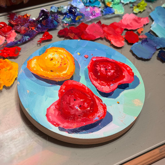 Original oil painting by Alai Ganuza titled "Heart madeleines". Three heart-shaped madeleine cakes, one golden orange and two berry red, arranged on a round light blue birch wood panel.