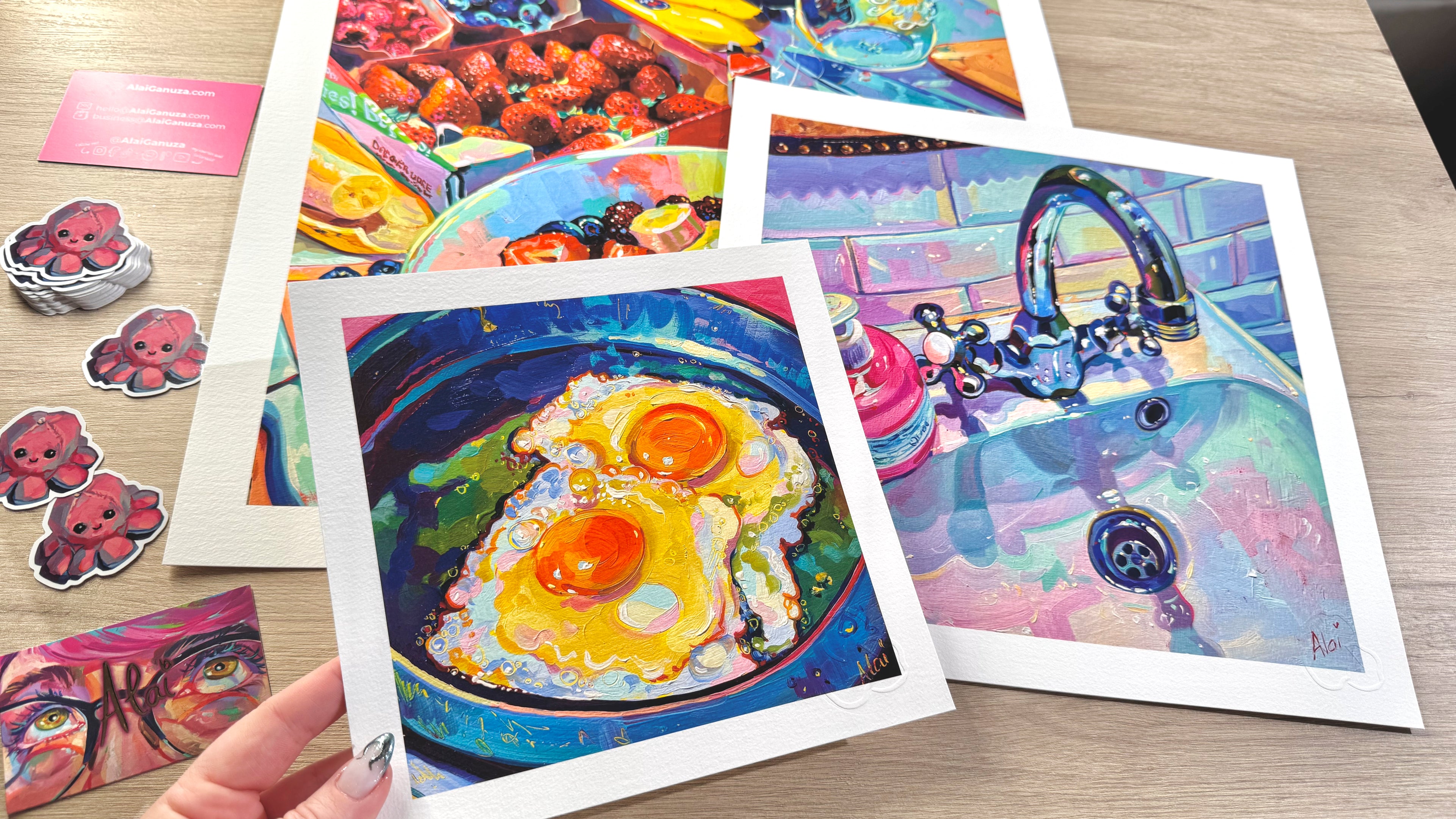 Collection of fine art prints by Alai Ganuza featuring colorful oil paintings of fried eggs, a bathroom sink, and a fruit bowl, displayed on a wooden table with stickers and cards.