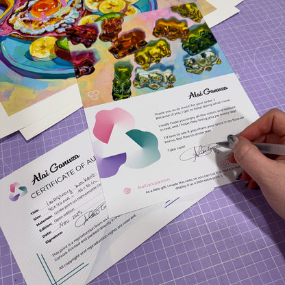 Hand holding a pen over a signed certificate of authenticity on a purple cutting mat with art prints in the background.
