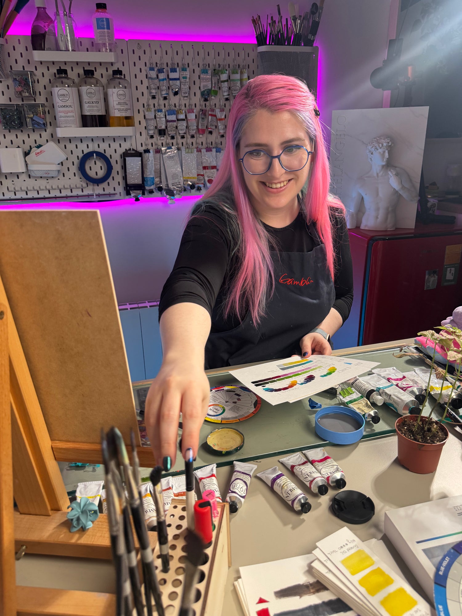 Artist Alai Ganuza in her studio, smiling while painting with oils and preparing her color palette, surrounded by brushes, art supplies, and warm studio lighting