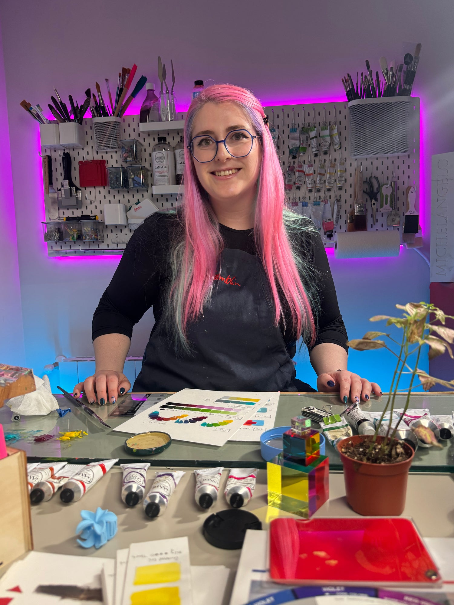 alai sitting in front of the camera, smilingin her studio, you can see all the brushes and paints laing around, she's wearing an apron and there are some color wheels in front of her, she made them as an excercise.
