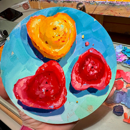 Original oil painting by Alai Ganuza titled "Heart madeleines". Three heart-shaped madeleine cakes, one golden orange and two berry red, arranged on a round light blue birch wood panel.