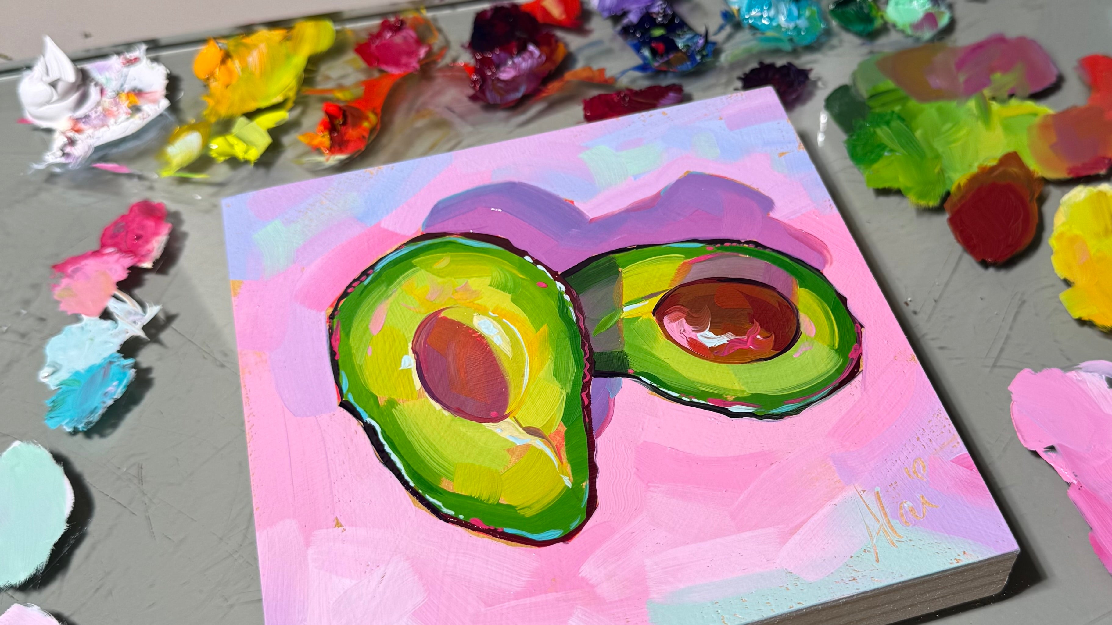 Colorful oil painting by Alai Ganuza depicting two avocado halves on a pink background, surrounded by a vibrant artist’s palette with mixed paints.