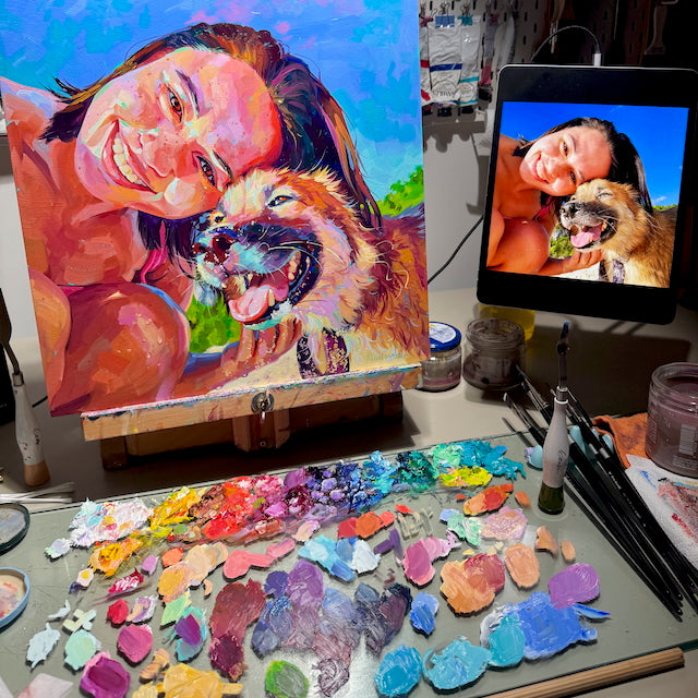 Custom oil painting commission by Alai Ganuza showing a woman and her dog smiling together, inspired by a reference photo displayed on a tablet beside the colorful paint palette.