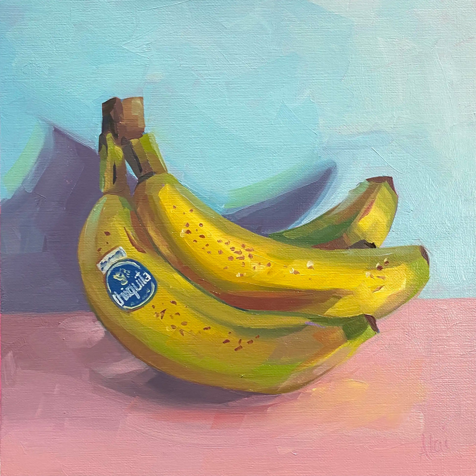 It depicts a vibrant still life study of a bunch of ripe yellow bananas resting on a soft pink surface against a pale blue background. The artwork features thick, blocky impasto oil brushstrokes on canvas, highlighting structured facets of lime green and golden ochre on the fruit peel, a prominent blue branding sticker, and a dramatic angular purple shadow casting behind them.