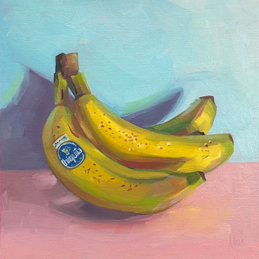 It depicts a vibrant still life study of a bunch of ripe yellow bananas resting on a soft pink surface against a pale blue background. The artwork features thick, blocky impasto oil brushstrokes on canvas, highlighting structured facets of lime green and golden ochre on the fruit peel, a prominent blue branding sticker, and a dramatic angular purple shadow casting behind them.