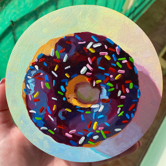 Original circular oil painting by Alai Ganuza titled "Chocolate sprinkle donut". A rich, dark-purple and chocolate glazed donut with vibrant multi-colored sprinkles, rendered in thick impasto on a soft yellow and blue background.