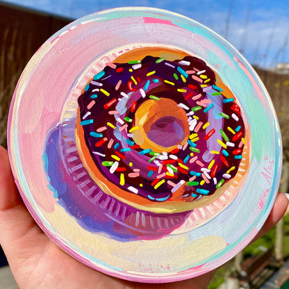 Original oil painting by Alai Ganuza titled "Rainbow Donut on Round". A donut with dark chocolate frosting and rainbow sprinkles painted on a circular wood panel against a swirling pastel pink and teal background.